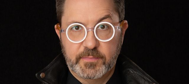 Close up headshot of artist Greg Wilder. Greg is wearing stylishly bright white circular glasses and a classic black leather jacket.